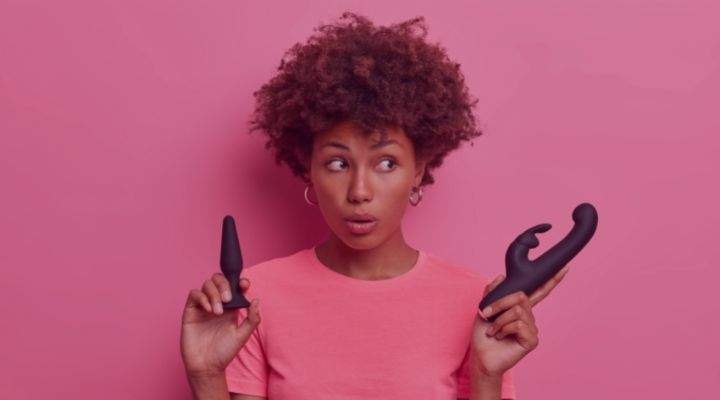 Imagem de uma mulher negra surpresa, olhando de lado e segurando vibrador e plug anal nas mãos.